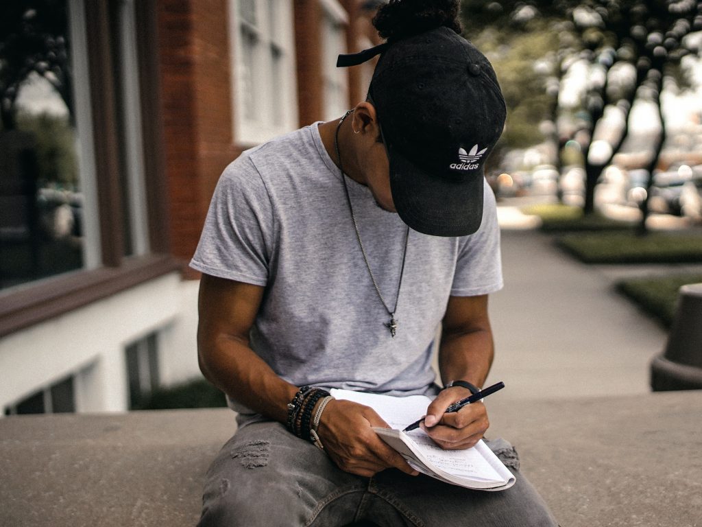Photo of a young man in a baseball cap sitting on a wall writing into a notebook